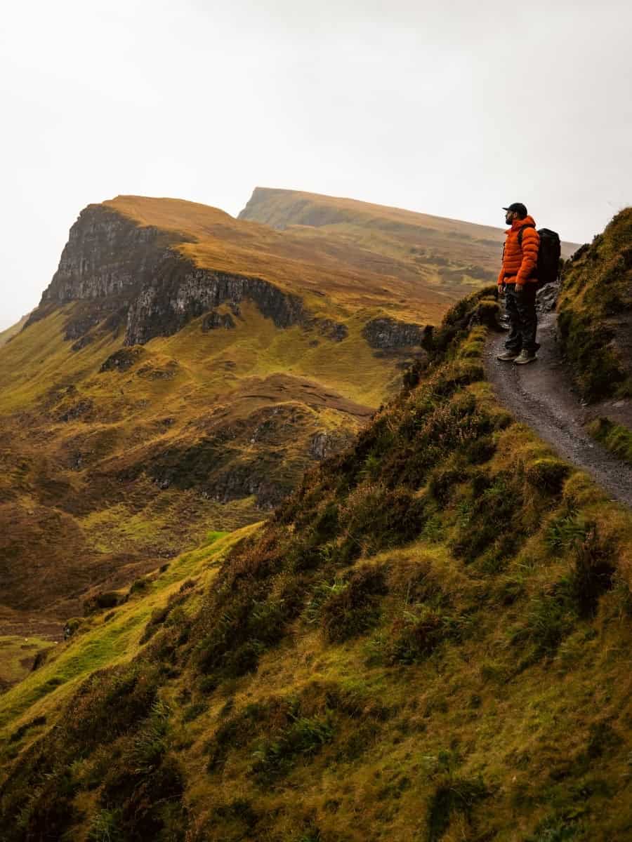 Wide landscape view of the Quiraing on the Isle of Skye with Joey standing in the distance among dramatic cliffs and rolling terrain.
