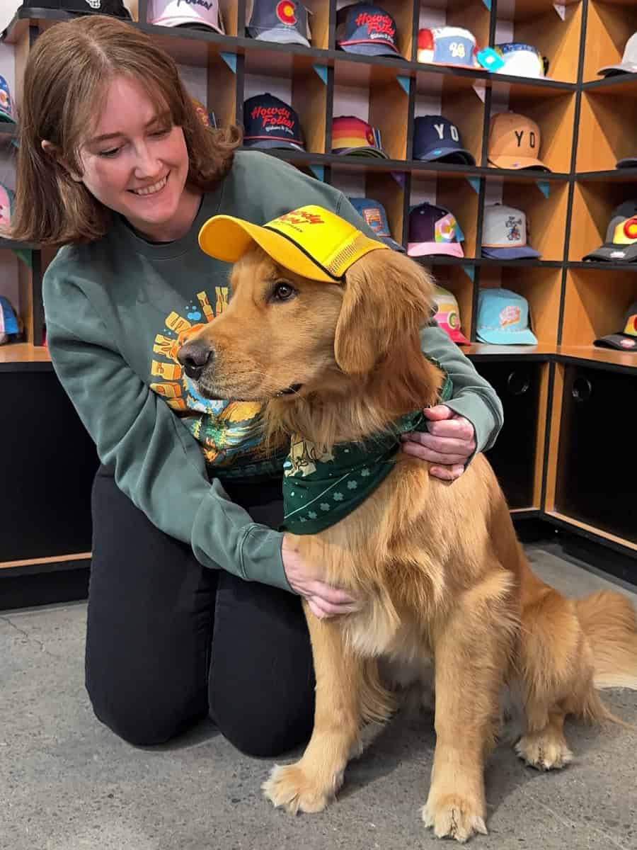 Katie sitting next to a dog named Champ wearing an official Goldens in Golden pup hat and bandana.
