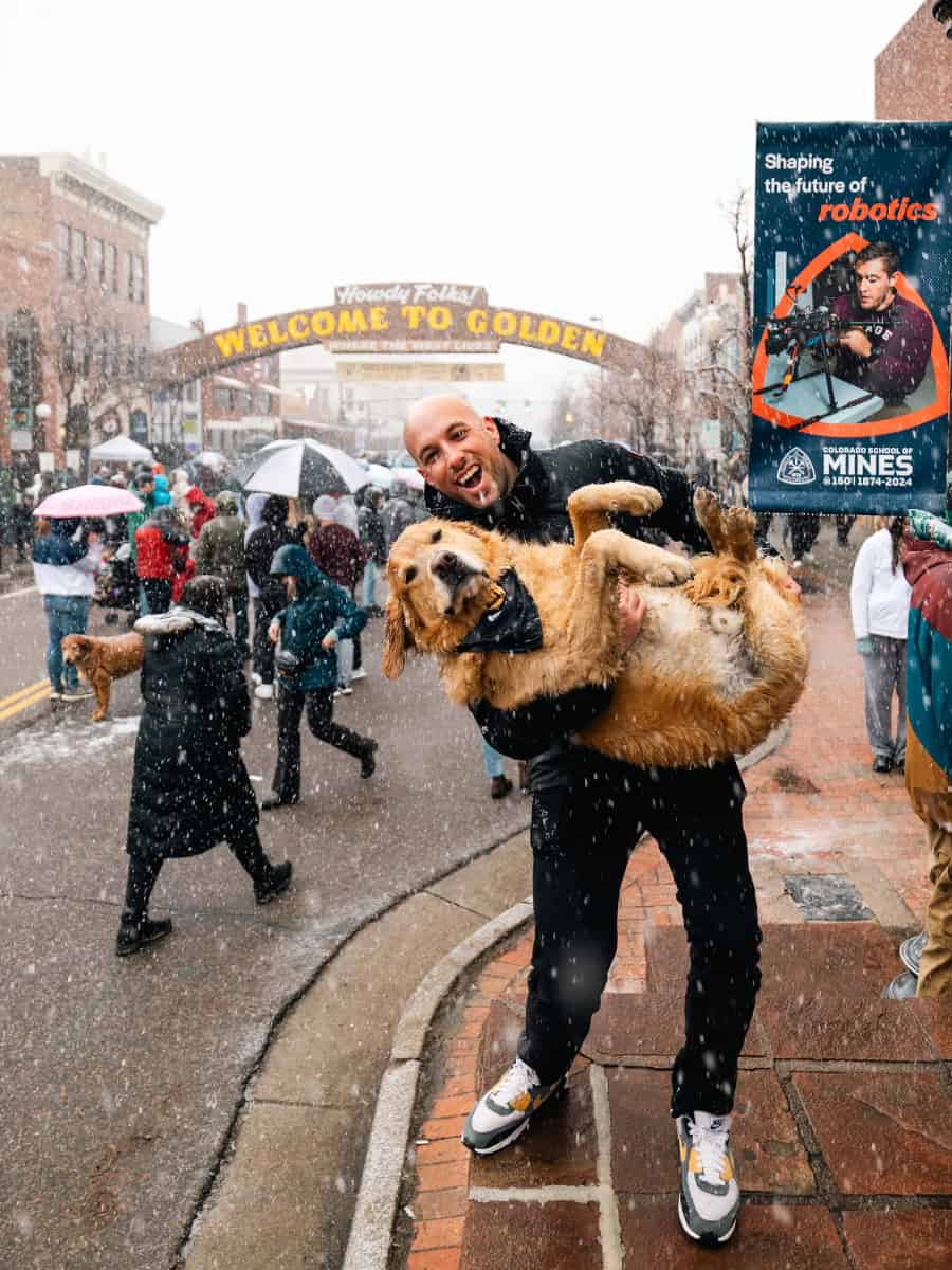 A man holding up his Golden Retriever in falling snow during the Goldens in Golden 2024 event in downtown Golden, Colorado.